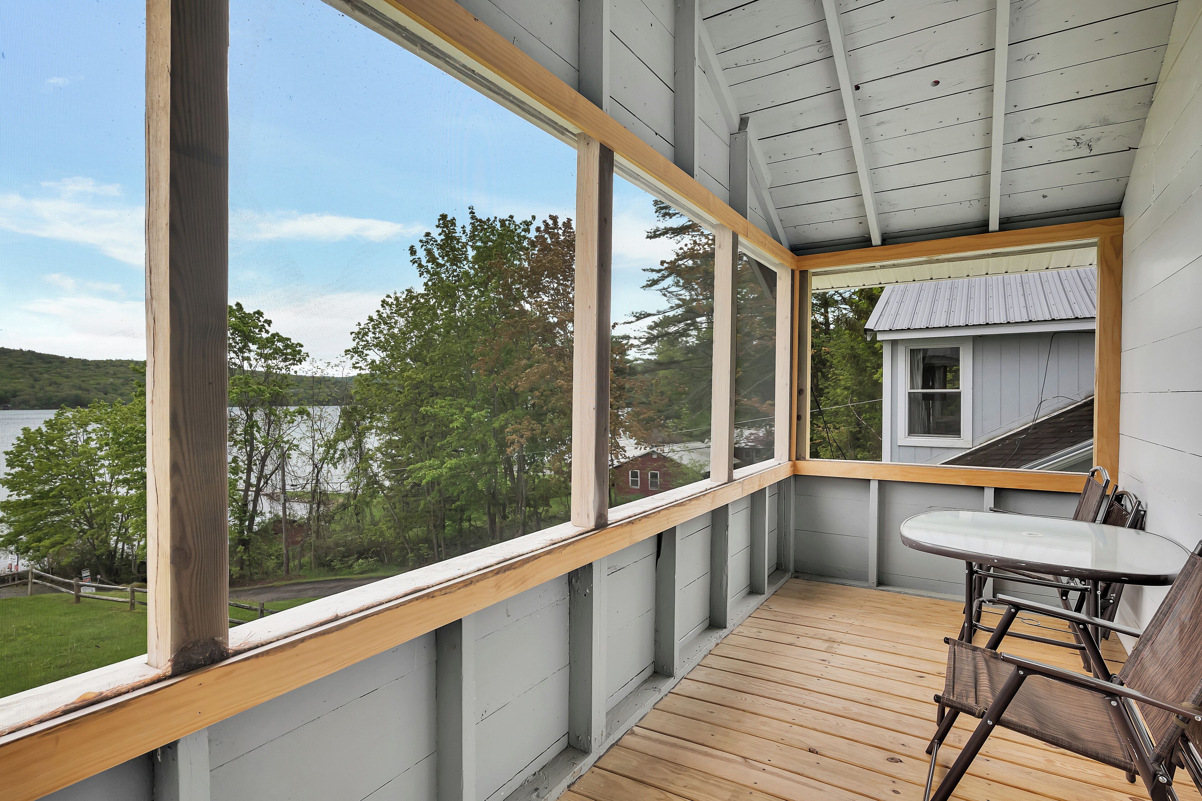 A covered porch with a wooden table and chairs, large windows offering a view of a lake and trees.