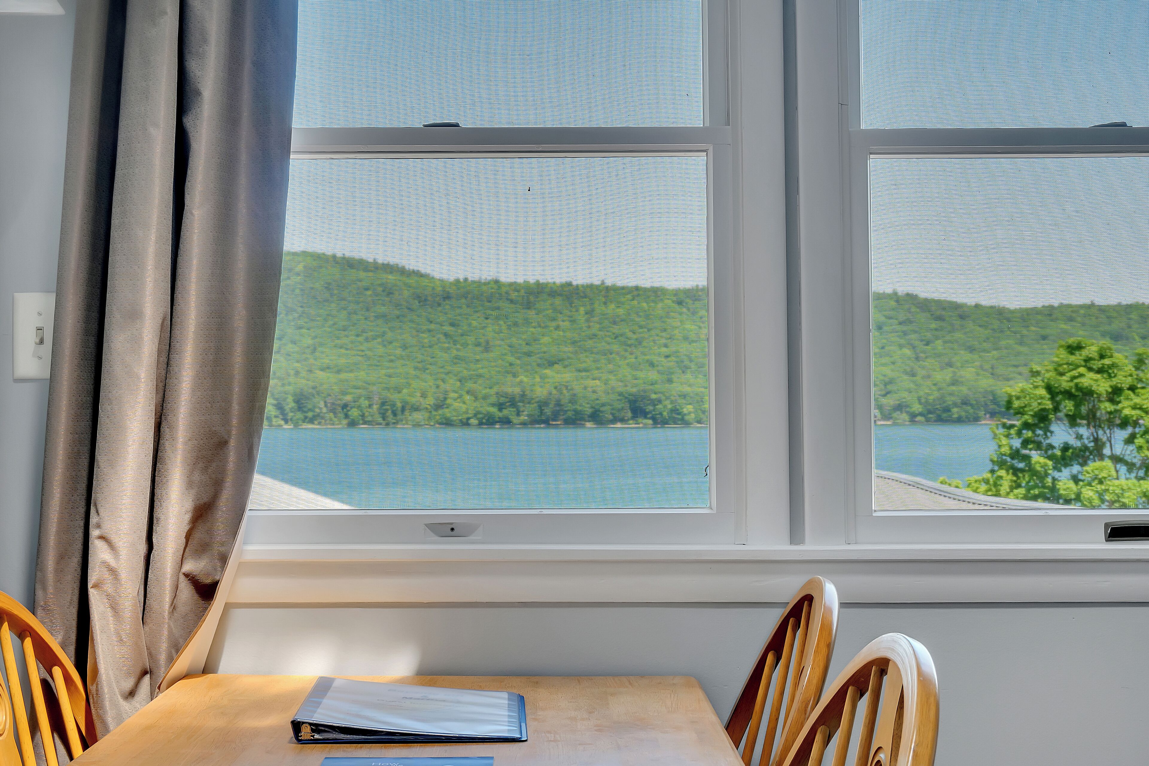 A room with a table and chairs, a window with a view of a lake and hills, and a curtain partially drawn.
