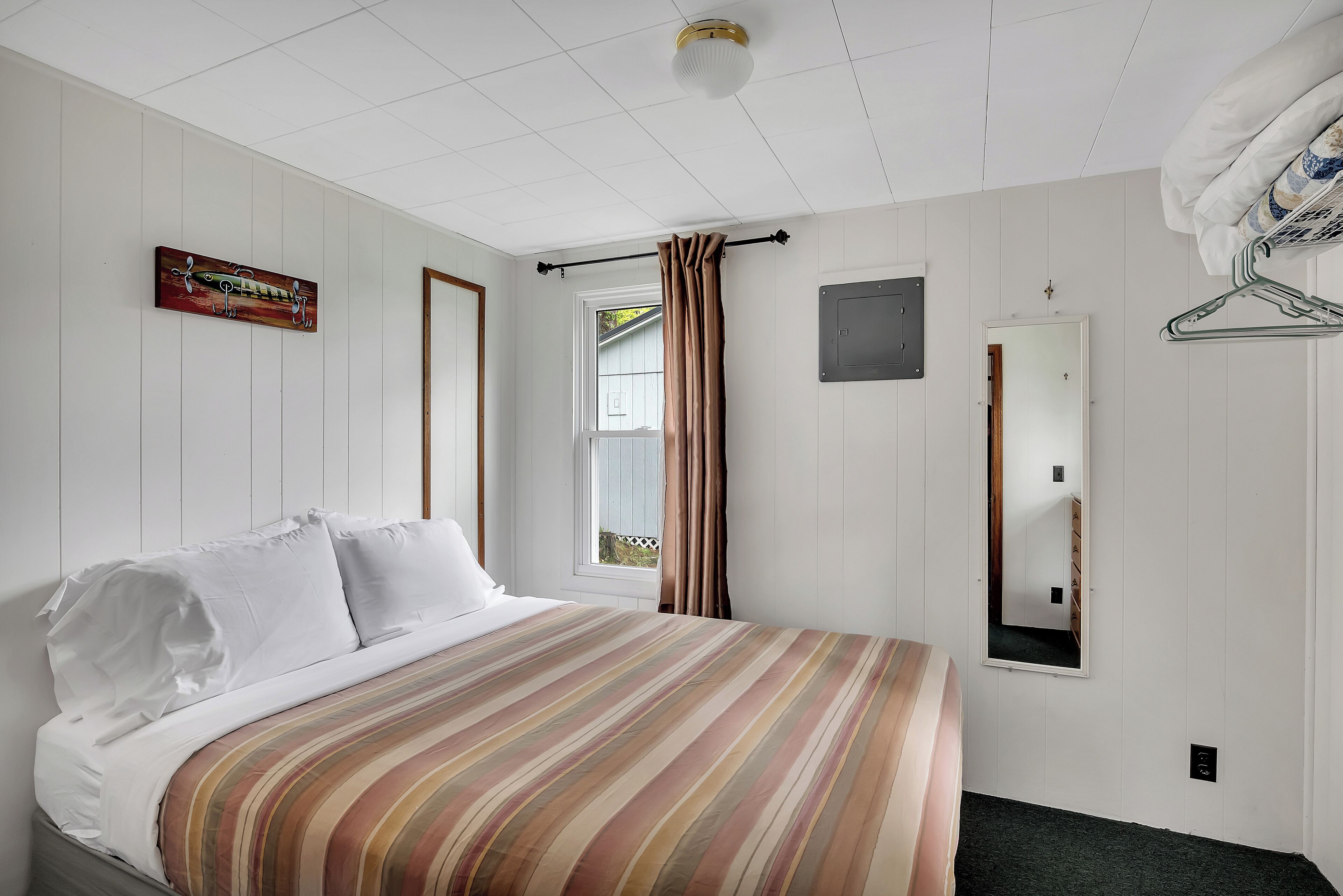 A hotel room with a bed, a window with curtains, a wall-mounted mirror, and a coat rack.