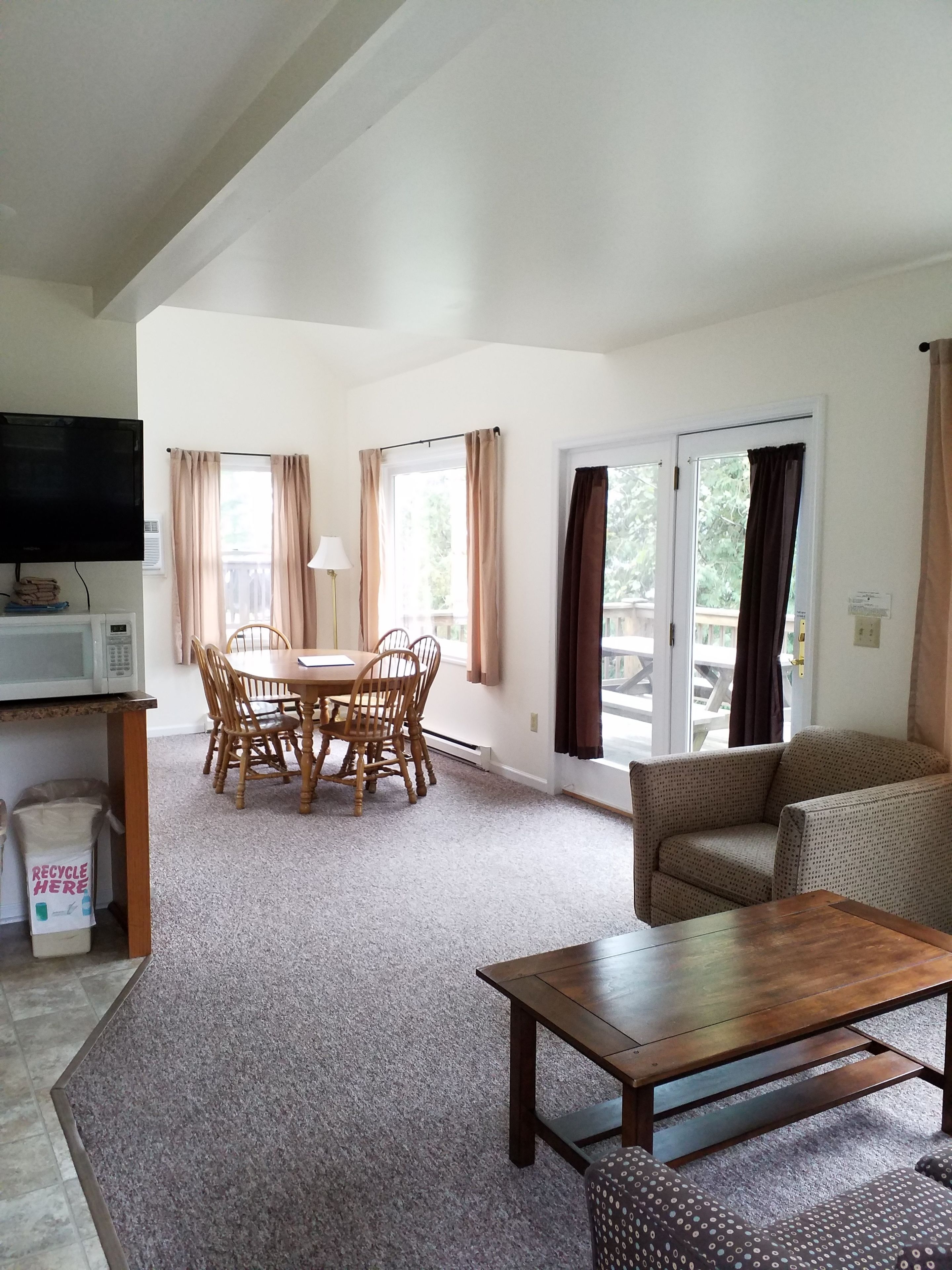 A living room with a wooden dining table and chairs, a sofa, a television, and a sliding glass door leading to an outdoor area.