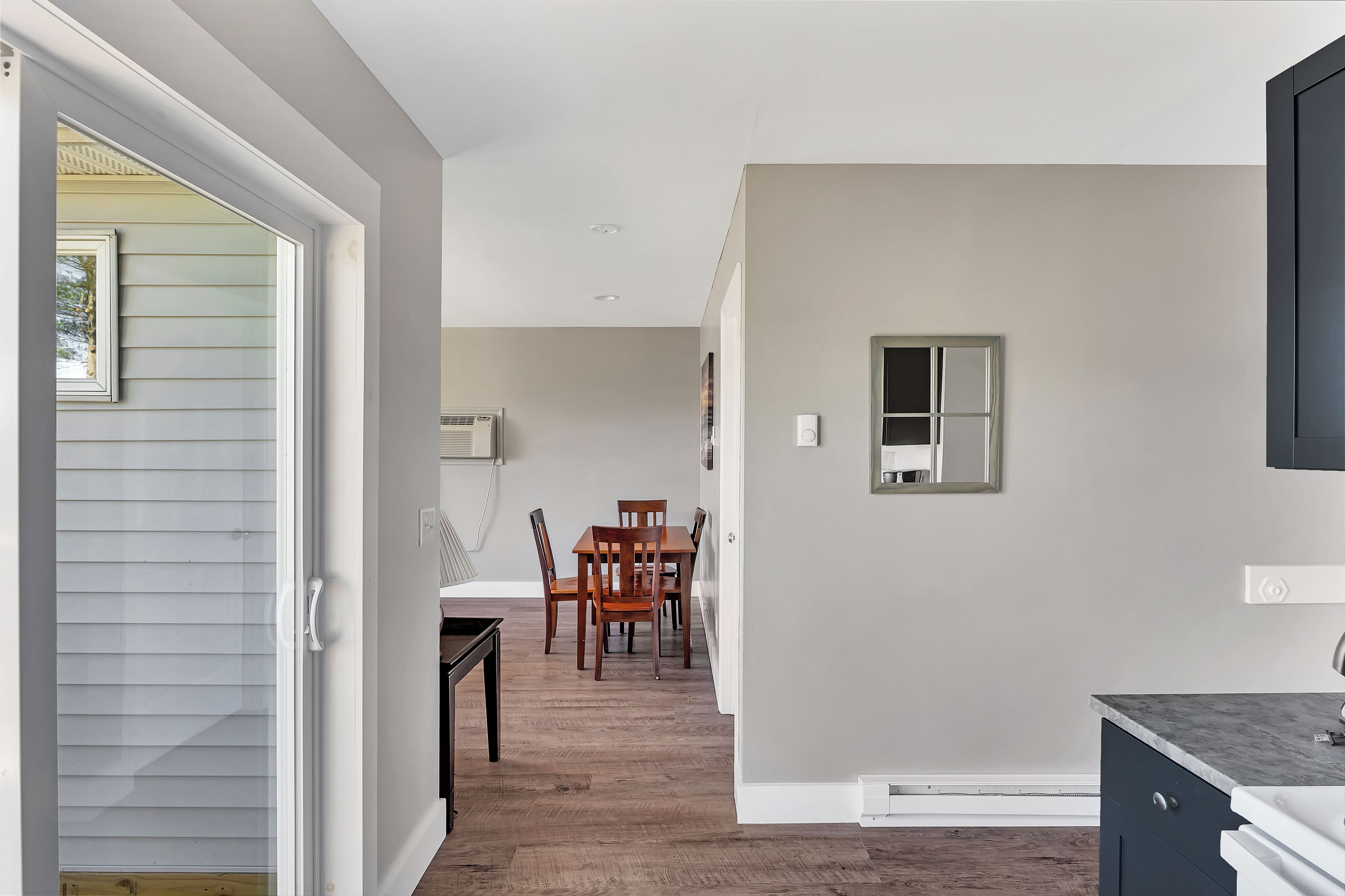 A modern kitchen with a dark countertop, a dining area with wooden chairs, and a hallway leading to a bedroom.