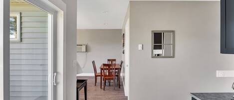 A modern kitchen with a dark countertop, a dining area with wooden chairs, and a hallway leading to a bedroom.