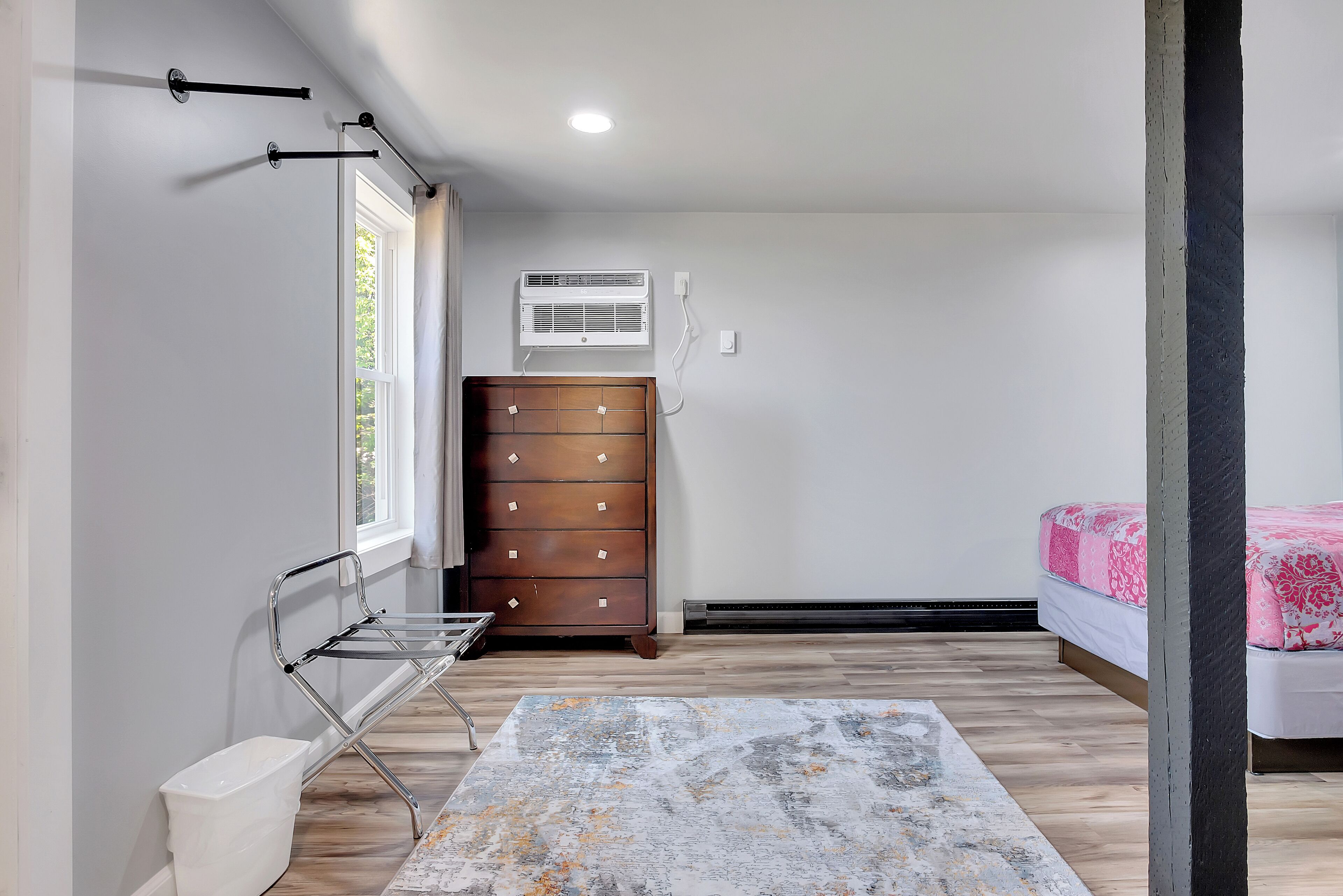Un dormitorio con un aparador de madera, una cama con ropa de cama rosa, una ventana con cortinas y una lámpara en el techo.