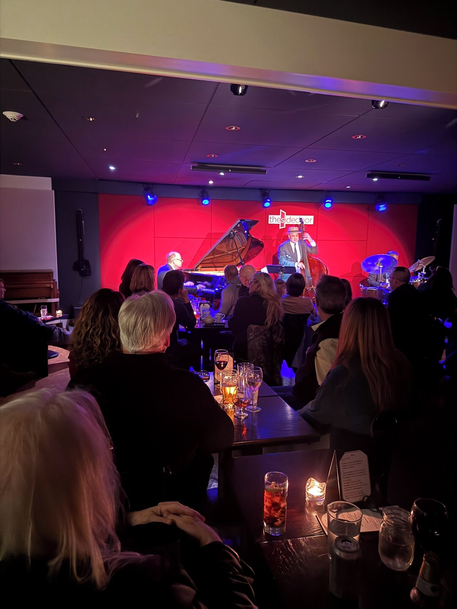 A concert with a grand piano and a band, seated audience, and a stage with a red backdrop.
