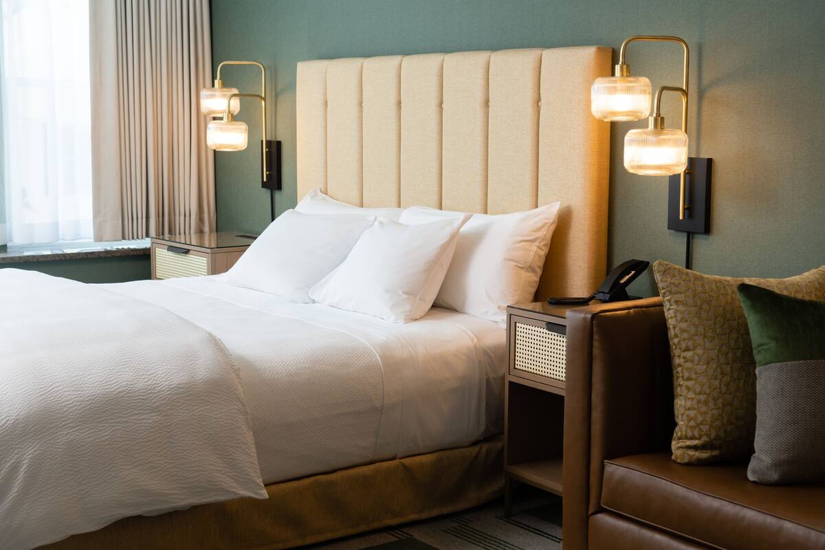 A neatly made bed with white linens, a brown leather sofa with cushions, and two wall-mounted lamps.