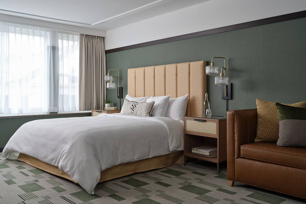 A neatly arranged hotel room with a large bed, a brown leather sofa, a nightstand with a lamp, and a view of the city through sheer curtains.