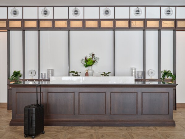 A reception desk with a white marble top, dark wood paneling, and a potted plant. A black suitcase is on the floor to the left.