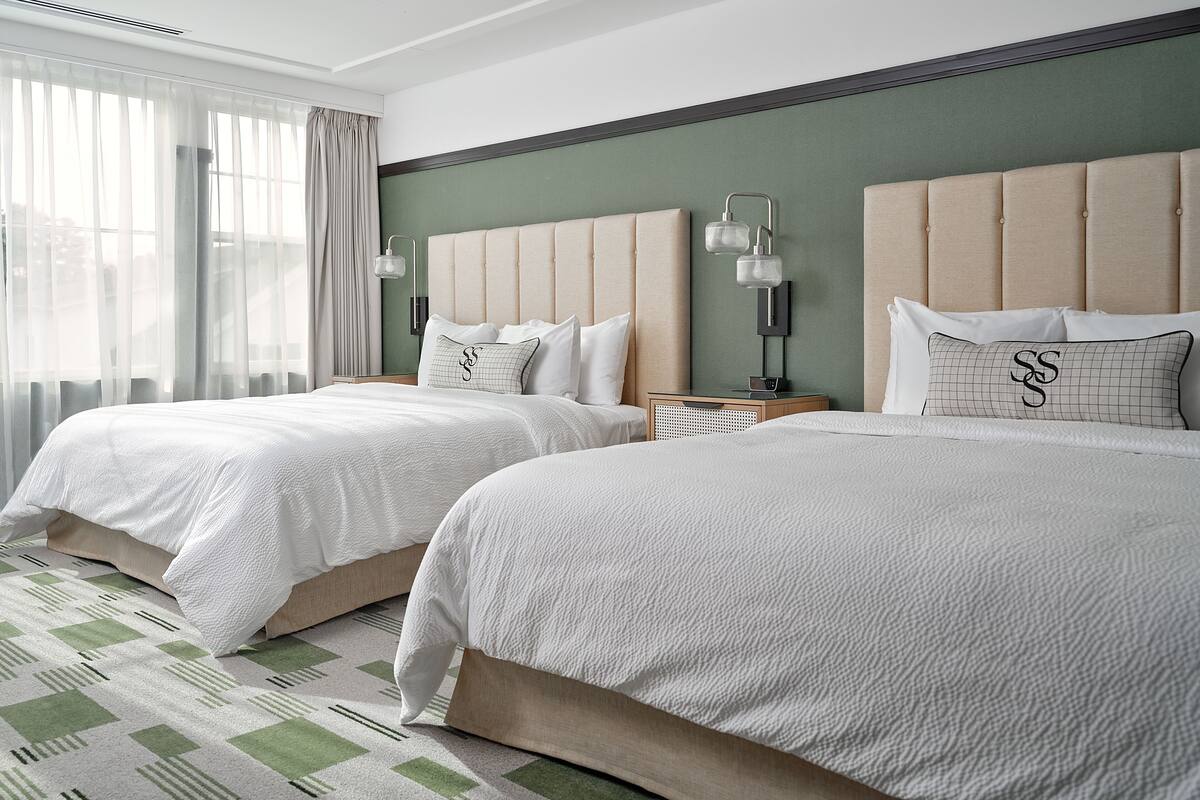 A hotel room with two beds, a green accent wall, and a large window with sheer curtains.