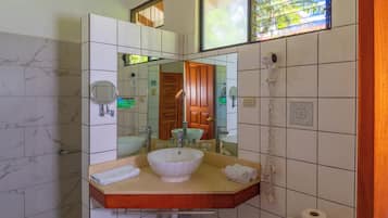 A bathroom with a marble-tiled wall, a wooden vanity with a sink, a mirror, a toilet, and a window with a view of greenery.