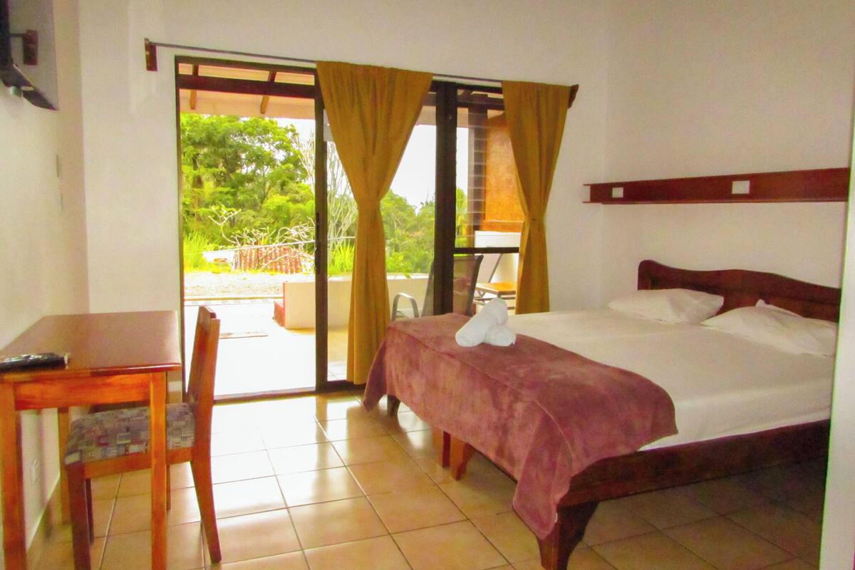 A hotel room with a bed, a desk, and a chair. There is a sliding glass door leading to an outdoor area.