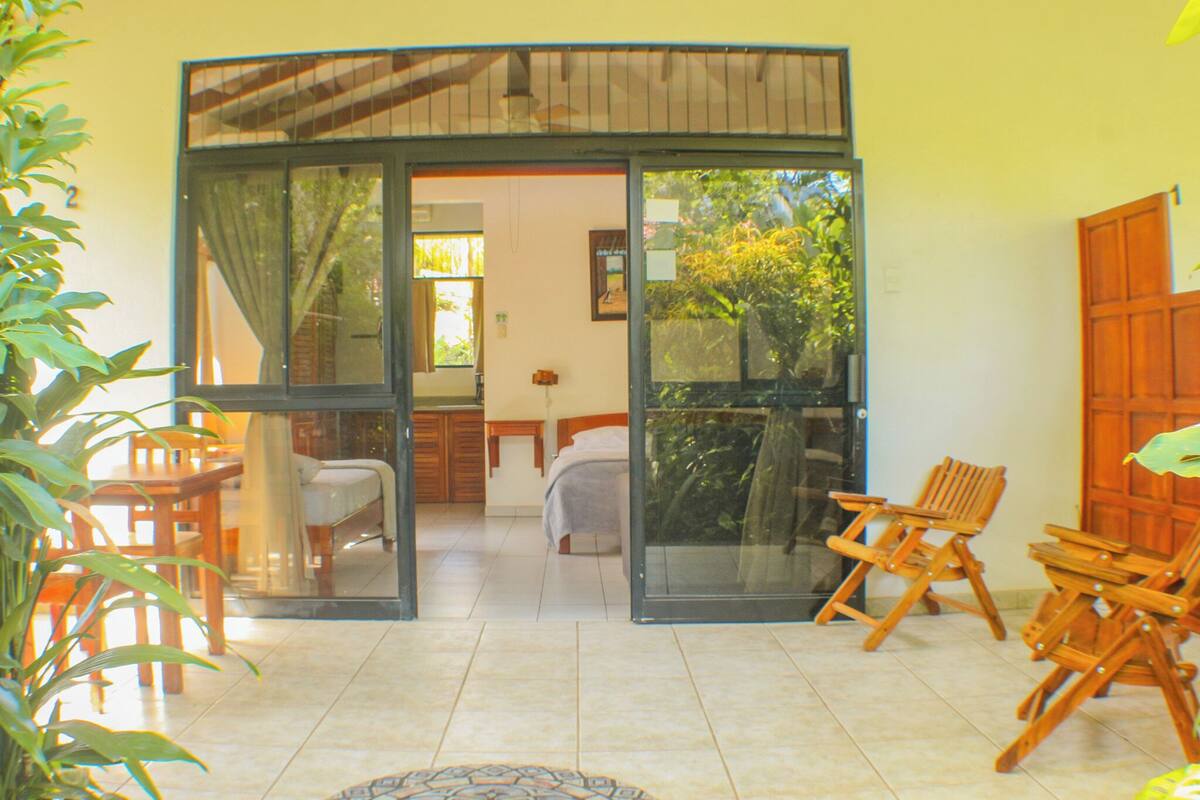 A room with a bed, a desk, and a chair, with a sliding glass door leading to an outdoor area with chairs and a table.