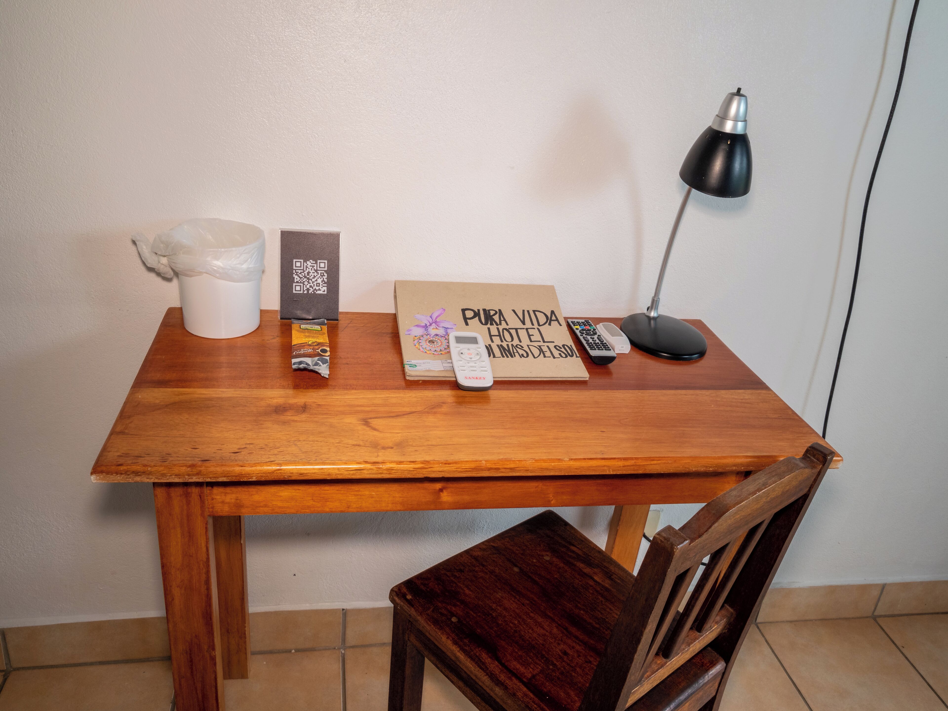 Une table en bois sur laquelle se trouvent un pichet blanc, une barre de chocolat, un sac portant un code QR, une enseigne d’hôtel, une télécommande, un téléphone et une lampe de bureau.