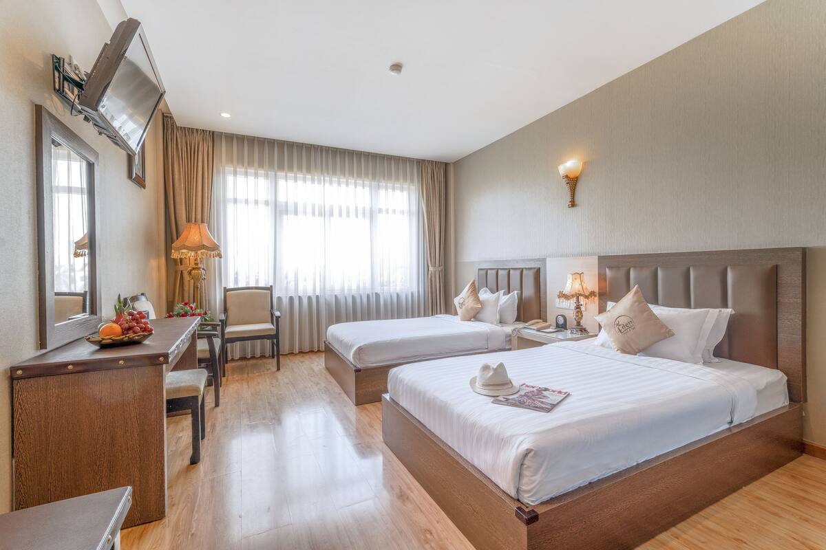 A hotel room with two beds, a desk, a chair, a lamp, and a television.