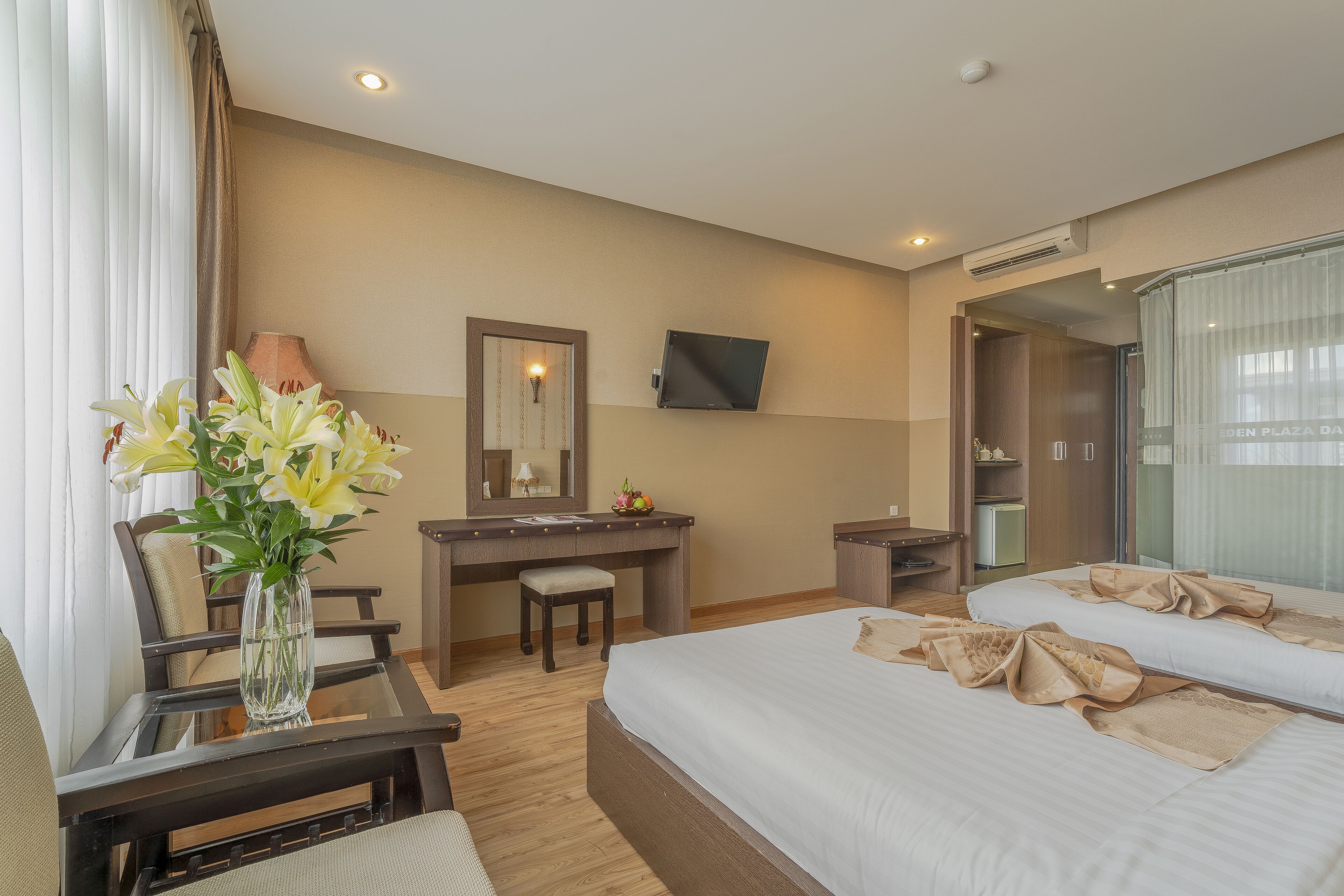 A hotel room with a bed, a desk, a chair, a vase of flowers, a mirror, and a television.