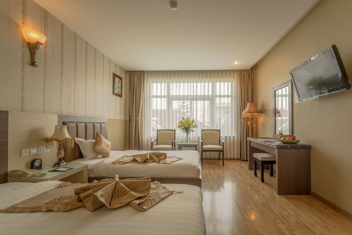 A hotel room with two beds, a desk with a lamp, a TV, and a window with curtains.