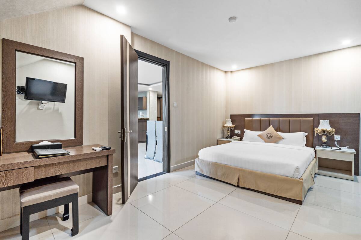 A hotel room with a bed, bedside tables, a desk with a chair, a television, and a door leading to another room.