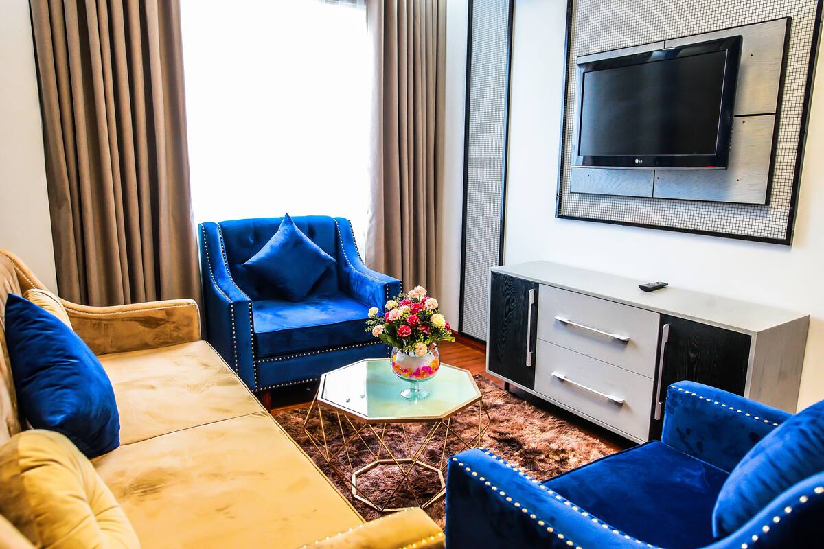 A living room with a sofa, two armchairs, a coffee table with flowers, a TV mounted on the wall, and a cabinet.