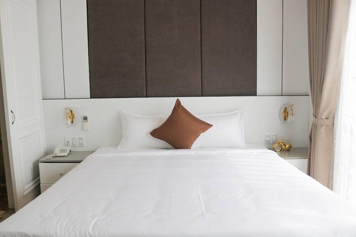 A neatly made bed with white linens and a single brown accent pillow, a bedside table with a phone and a small decorative item, and a wall-mounted light fixture.