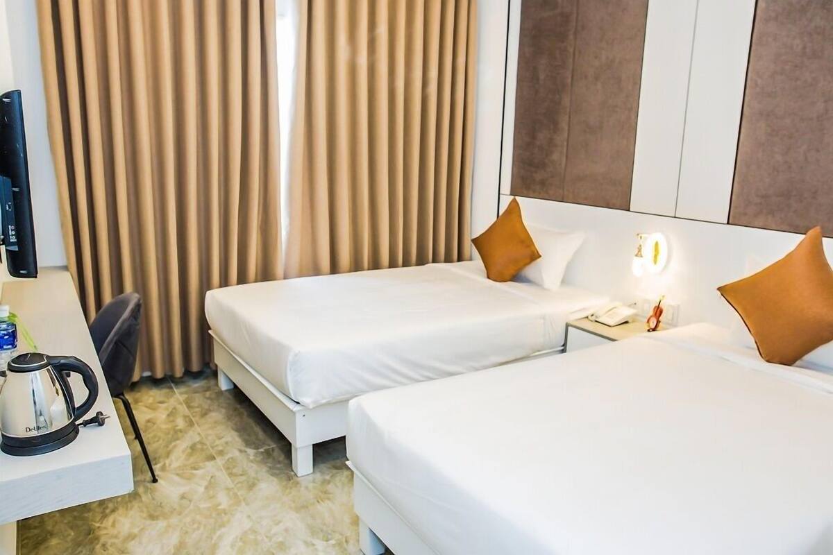 A hotel room with two beds, a desk with a kettle, a chair, and a curtain.