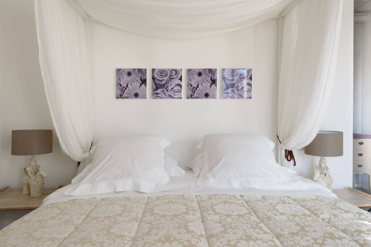 A bedroom with a canopy bed, two bedside tables with lamps, and three wall-mounted floral paintings.