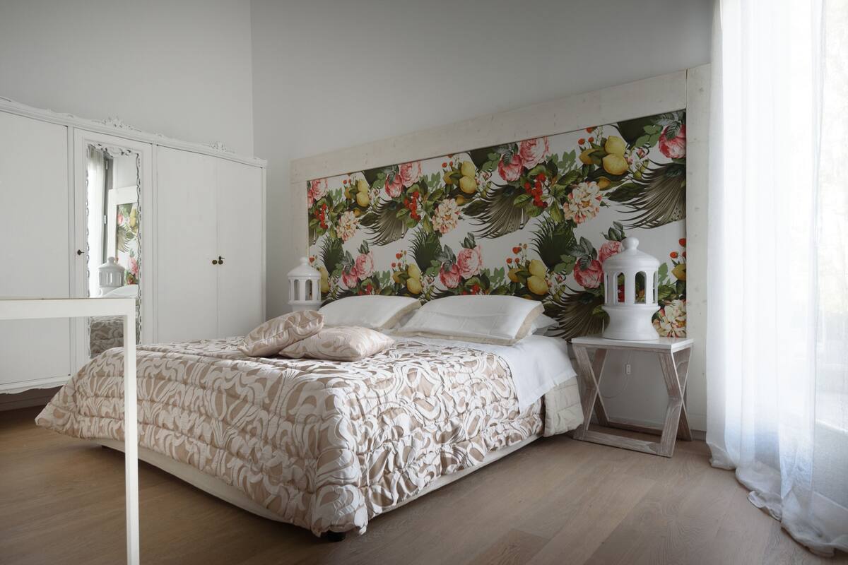 A bedroom with a bed, white nightstands, a mirror, and a floral wallpaper.