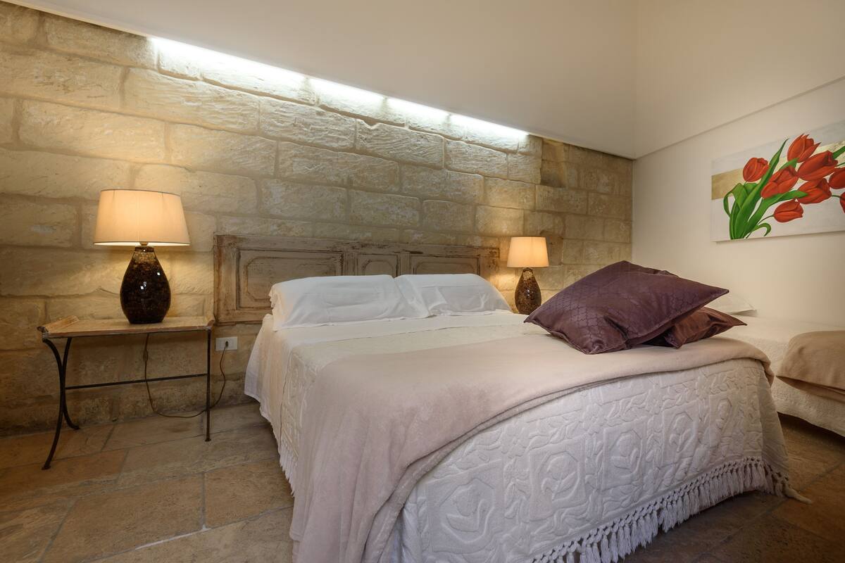 A bedroom with a stone wall, a bed with a white bedspread and a purple pillow, two bedside tables with lamps, and a painting of tulips on the wall.