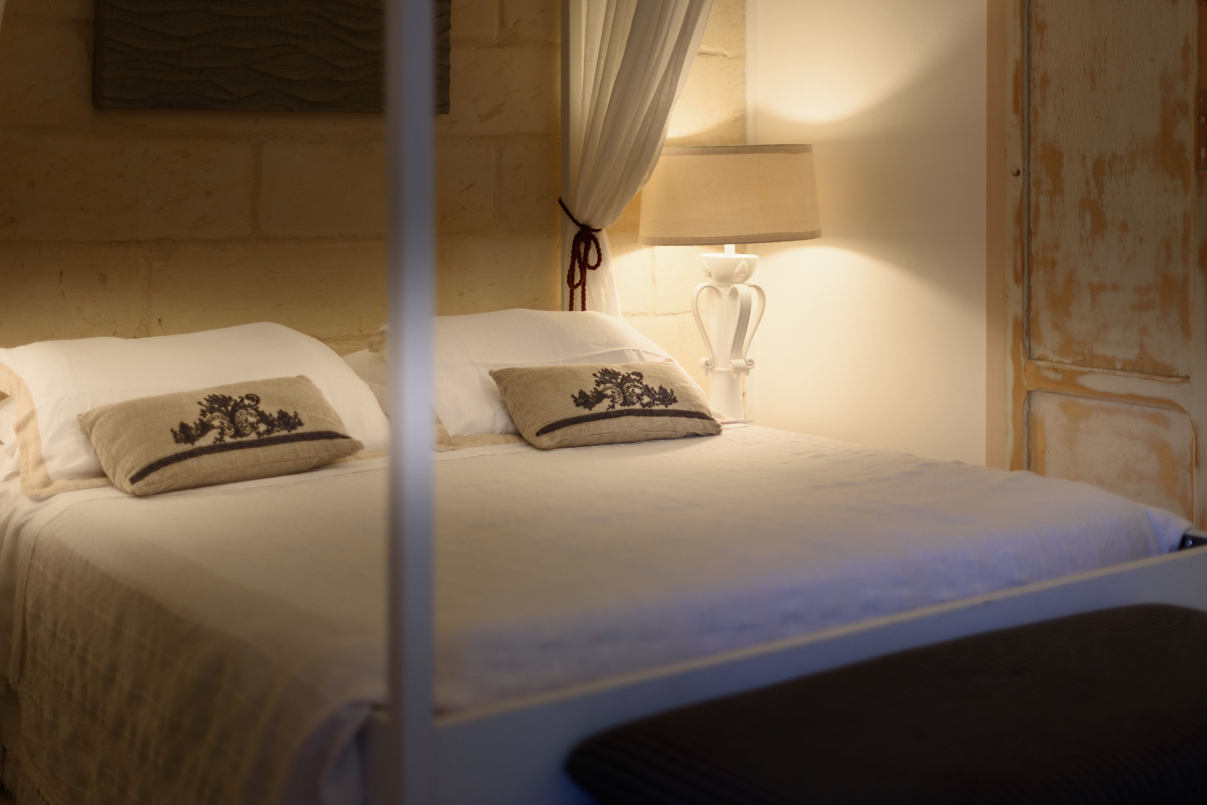 A neatly made bed with white linens and decorative pillows, a bedside lamp, and a curtain.