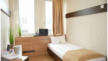 Una habitación de hotel pequeña con una cama individual, un escritorio con silla, una planta en maceta y un ventanal con cortinas.