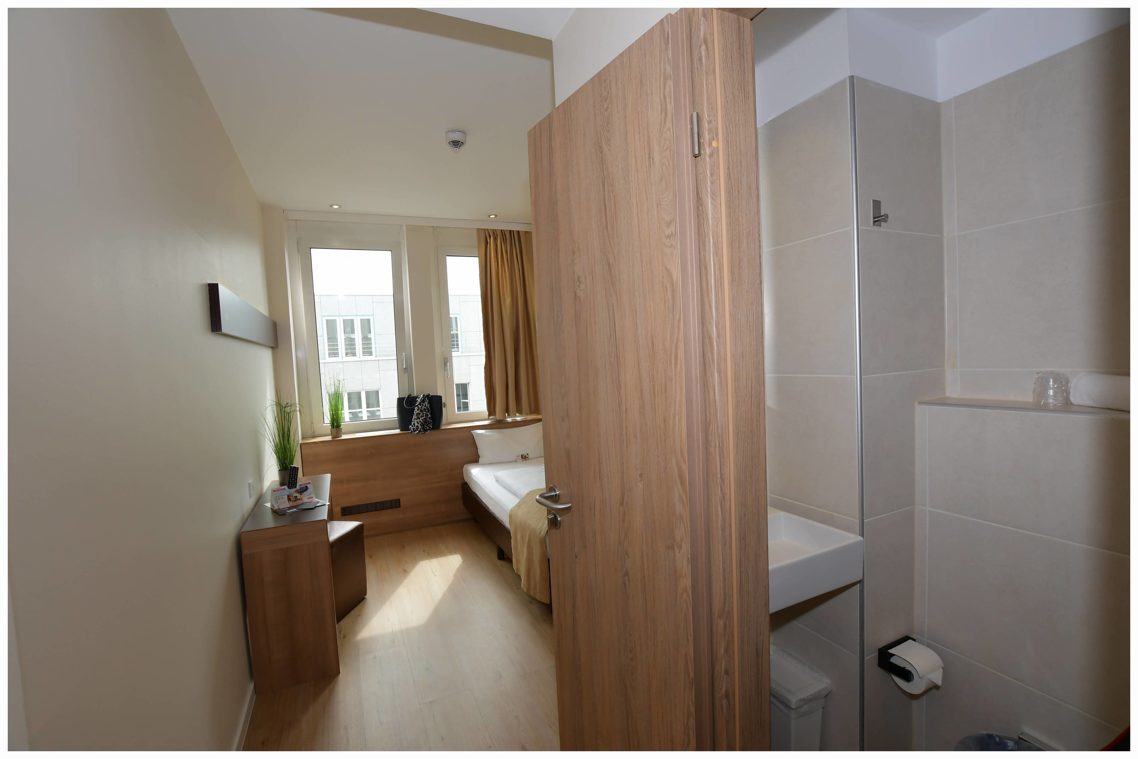 Un baño moderno con ducha, inodoro y lavamanos. Hay una ventana con cortinas y una pequeña zona de estar con un banco y una planta.