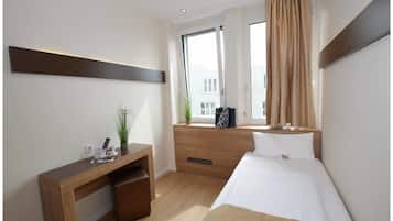 A hotel room with a bed, a desk, a window with curtains, a potted plant, and a view of buildings outside.