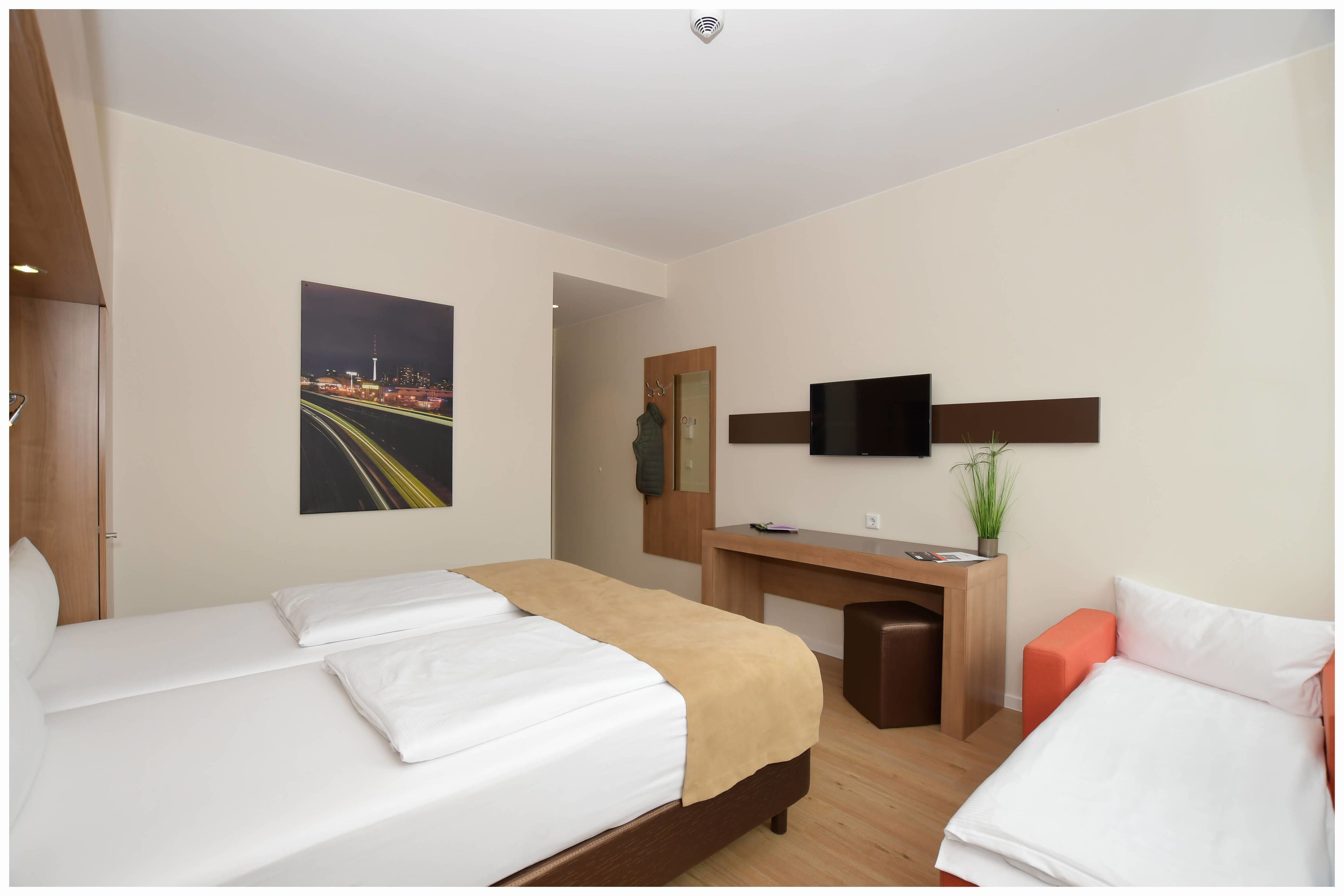 A hotel room with two beds, a desk, a TV, and a painting of a cityscape.