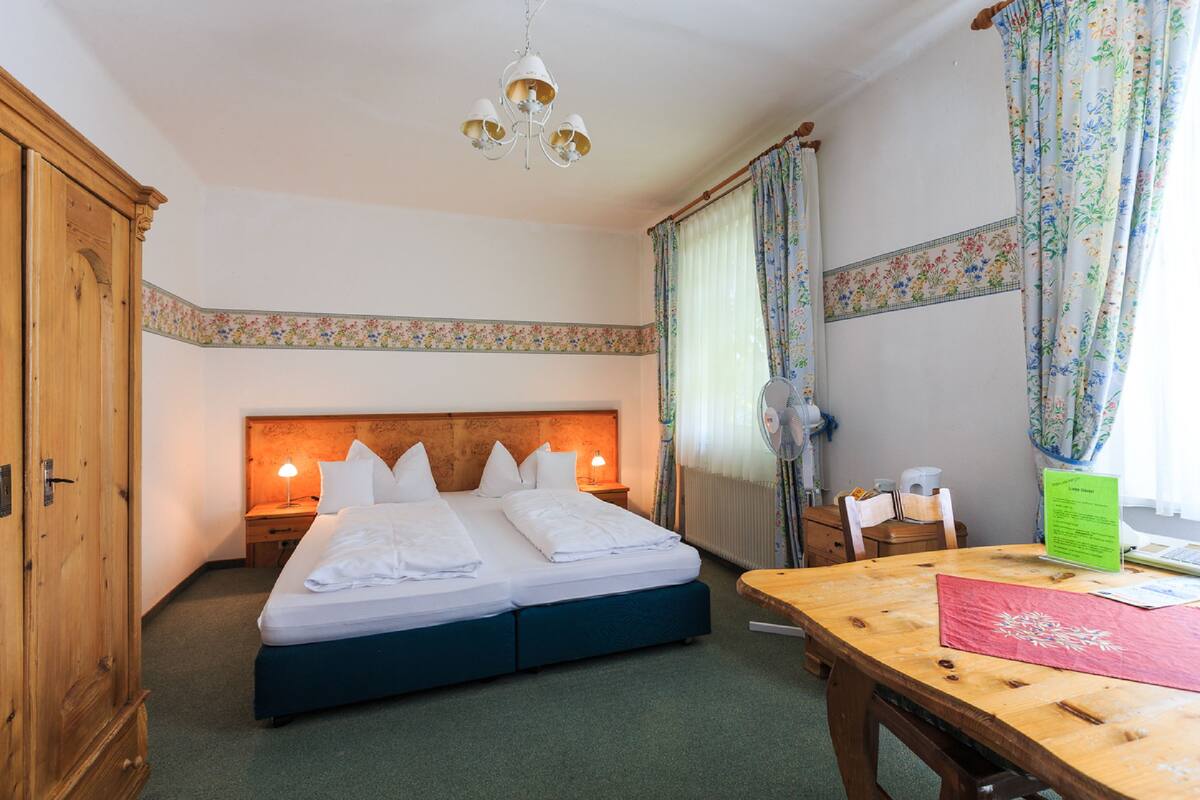 A hotel room with a double bed, wooden headboard, two bedside tables with lamps, a wooden desk, a chair, and a radiator.