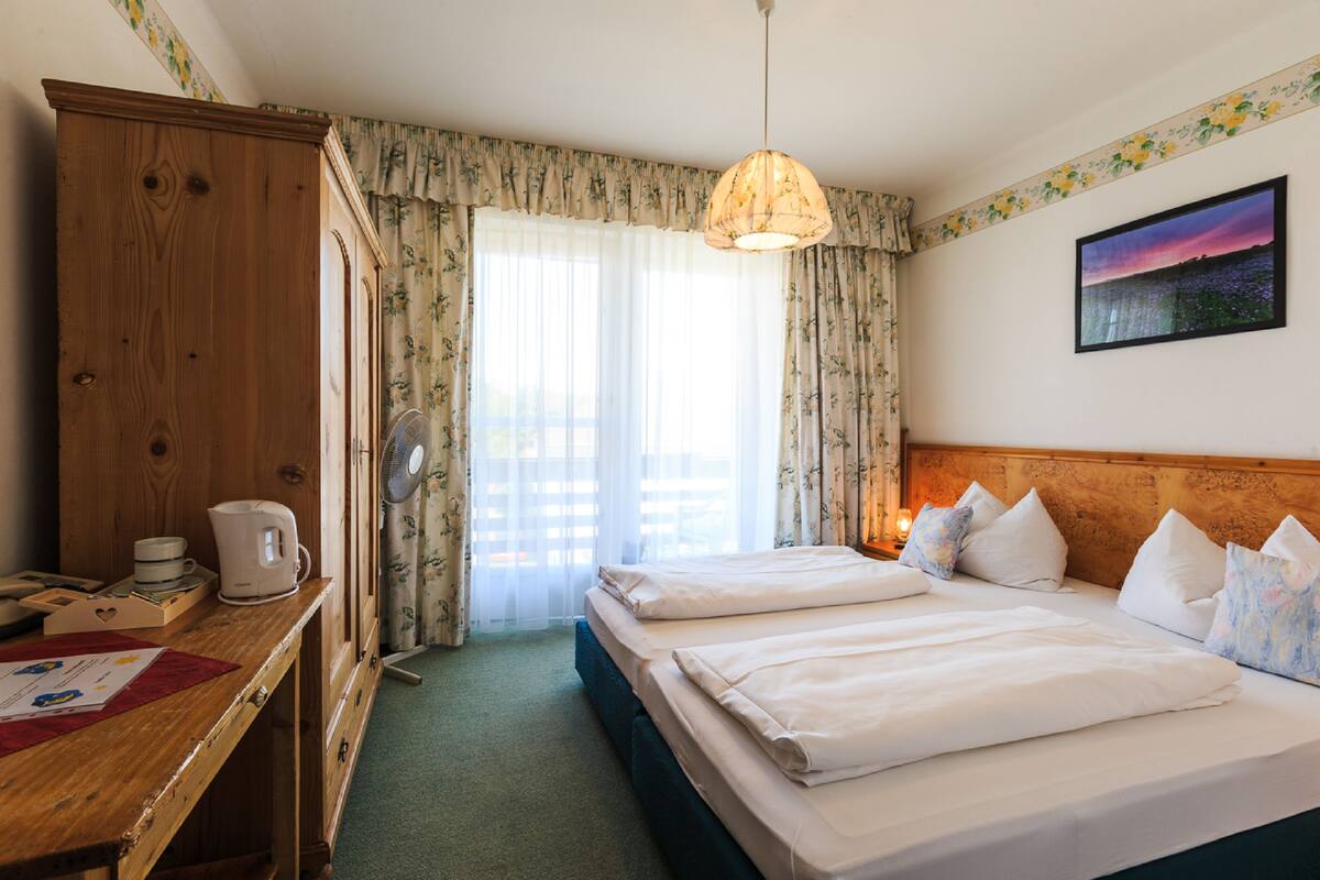 A hotel room with a wooden wardrobe, a desk with a kettle, two beds with white linens, a ceiling fan, and a painting on the wall.