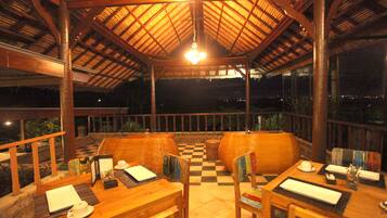 A covered outdoor dining area with wooden tables and chairs, a checkered floor, and a thatched roof.