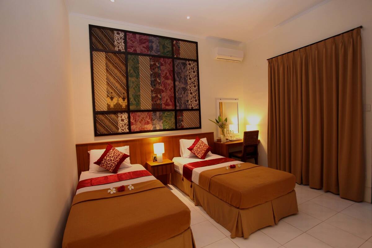A hotel room with two beds, a large decorative wall piece, a desk with a lamp, and a window with curtains.