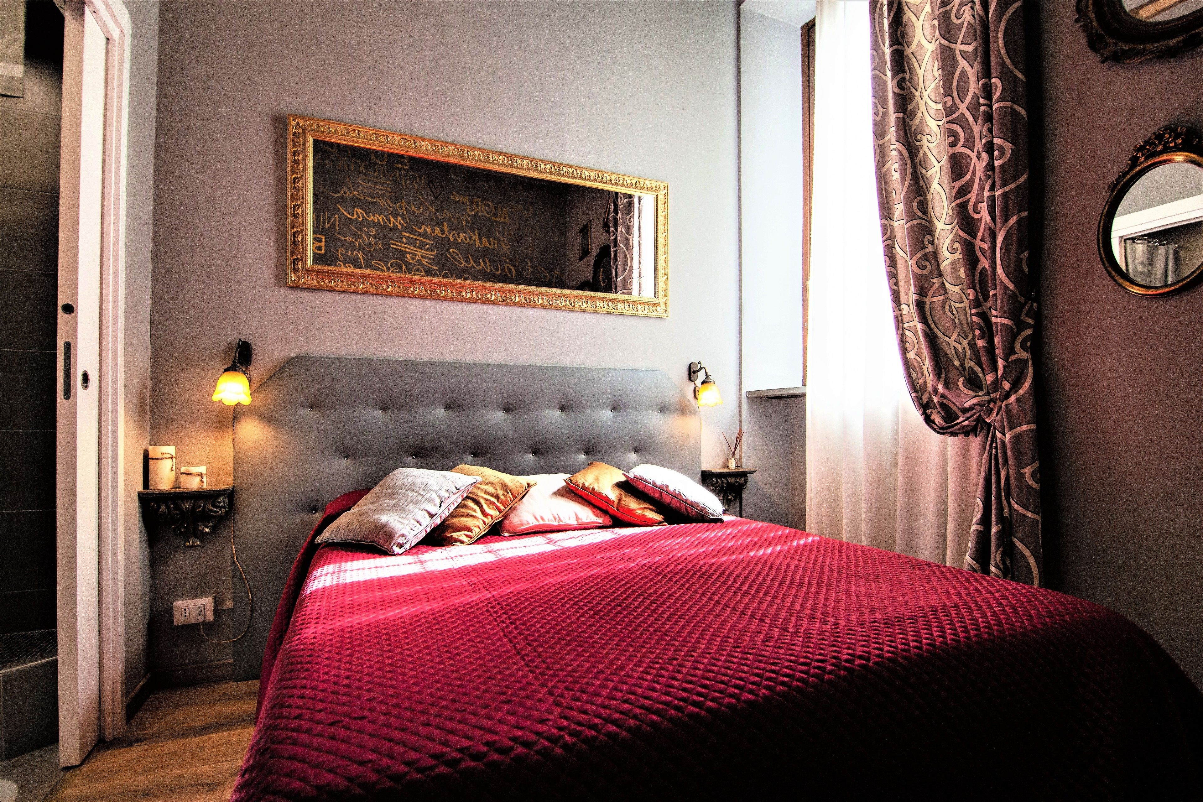 A bedroom with a tufted headboard, a large mirror, a bedside table with a lamp, and a red bedspread.