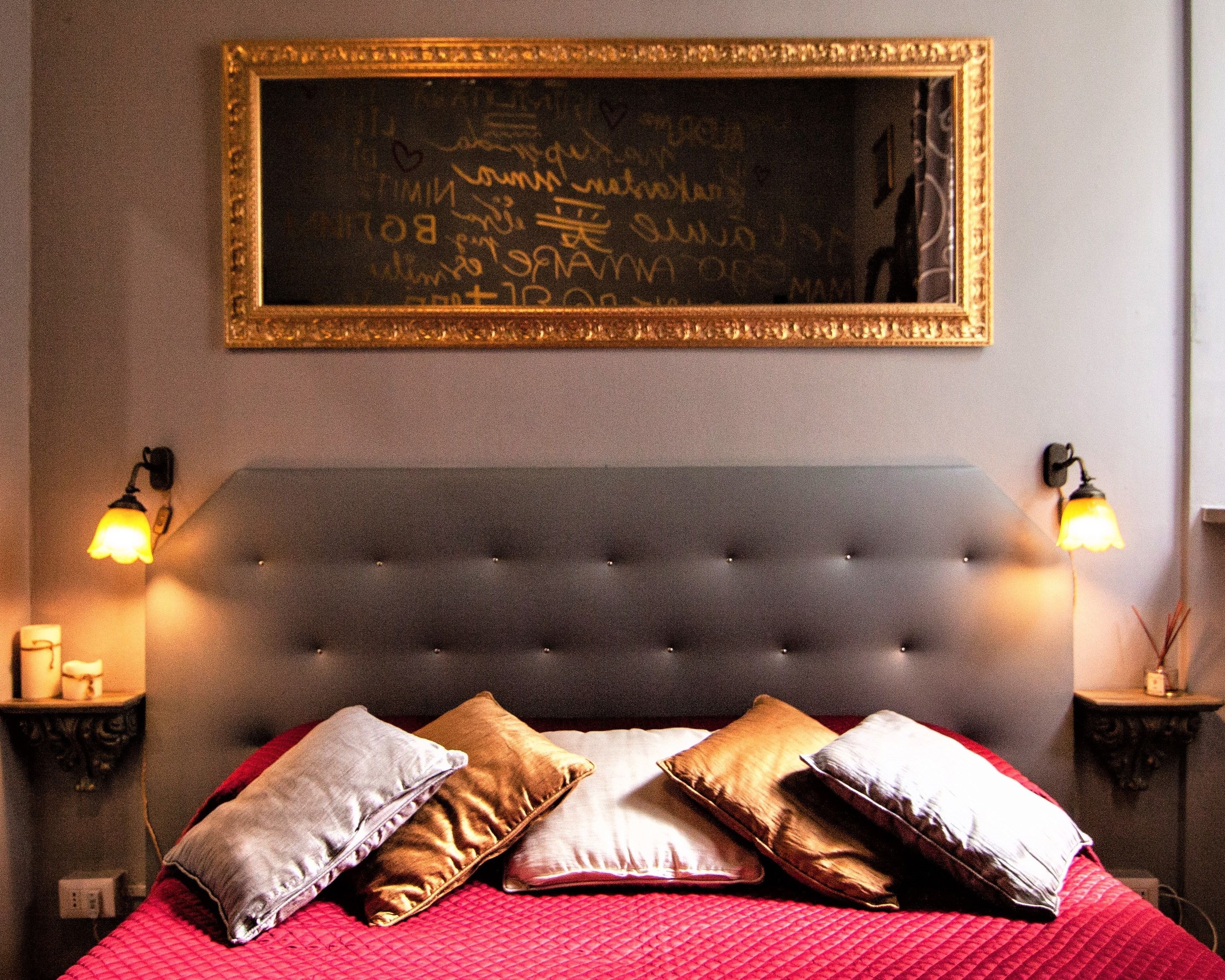 Une chambre à coucher avec une tête de lit capitonnée, une couvre-lit rouge et plusieurs coussins. Un tableau encadré est accroché au mur, ainsi que deux lampes fixées au mur.
