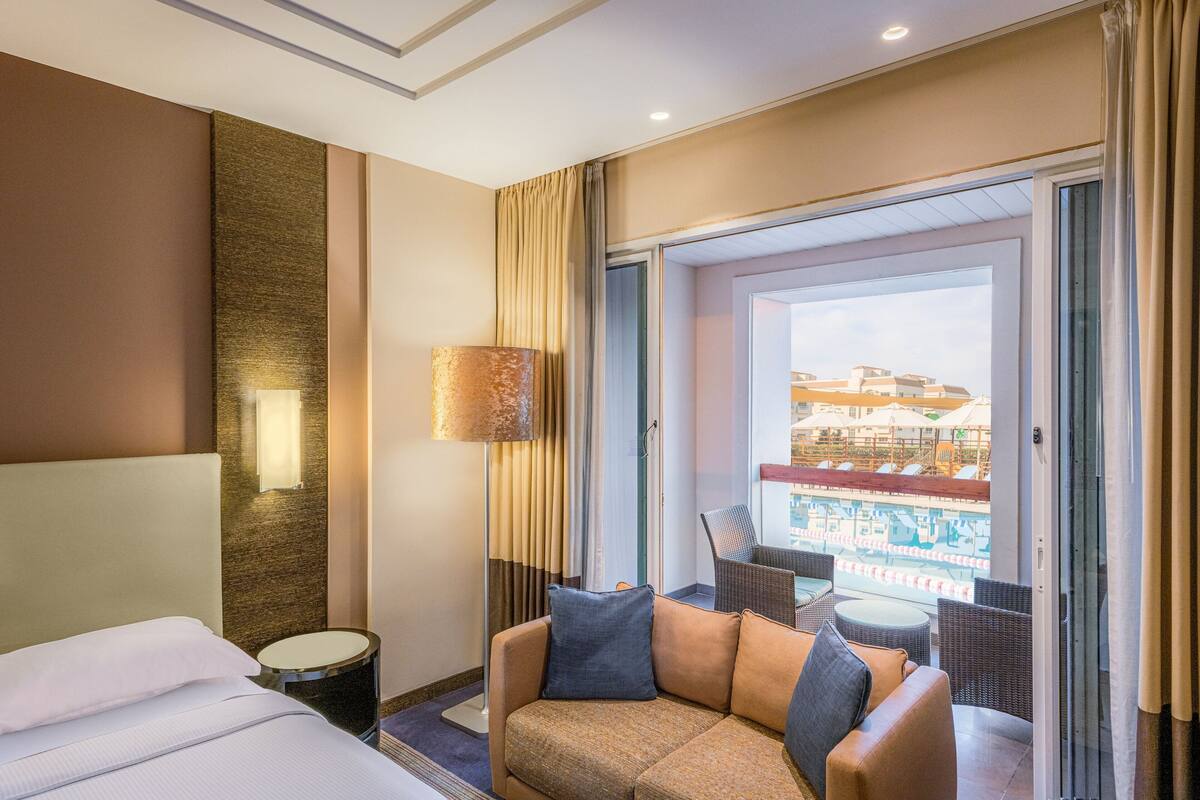 A hotel room with a bed, a sofa, a small table, and a balcony with a view of buildings.