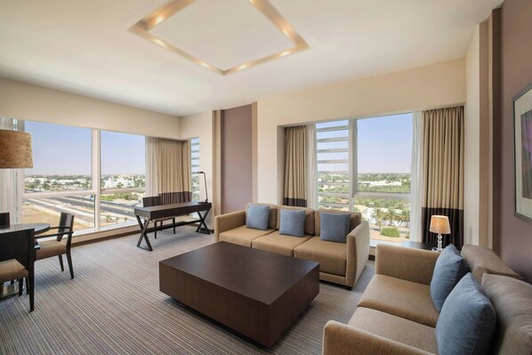 A spacious hotel room with a large window offering a city view, a sofa set, a coffee table, and a desk with a lamp. - Four Points by Sheraton Al Ain (Al Ain)