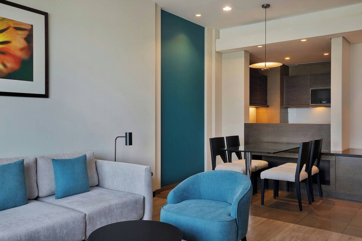 A modern living room with a grey sofa, a blue armchair, and a black coffee table. There is a dining area with a table and chairs, and a kitchen area with cabinets and a TV.