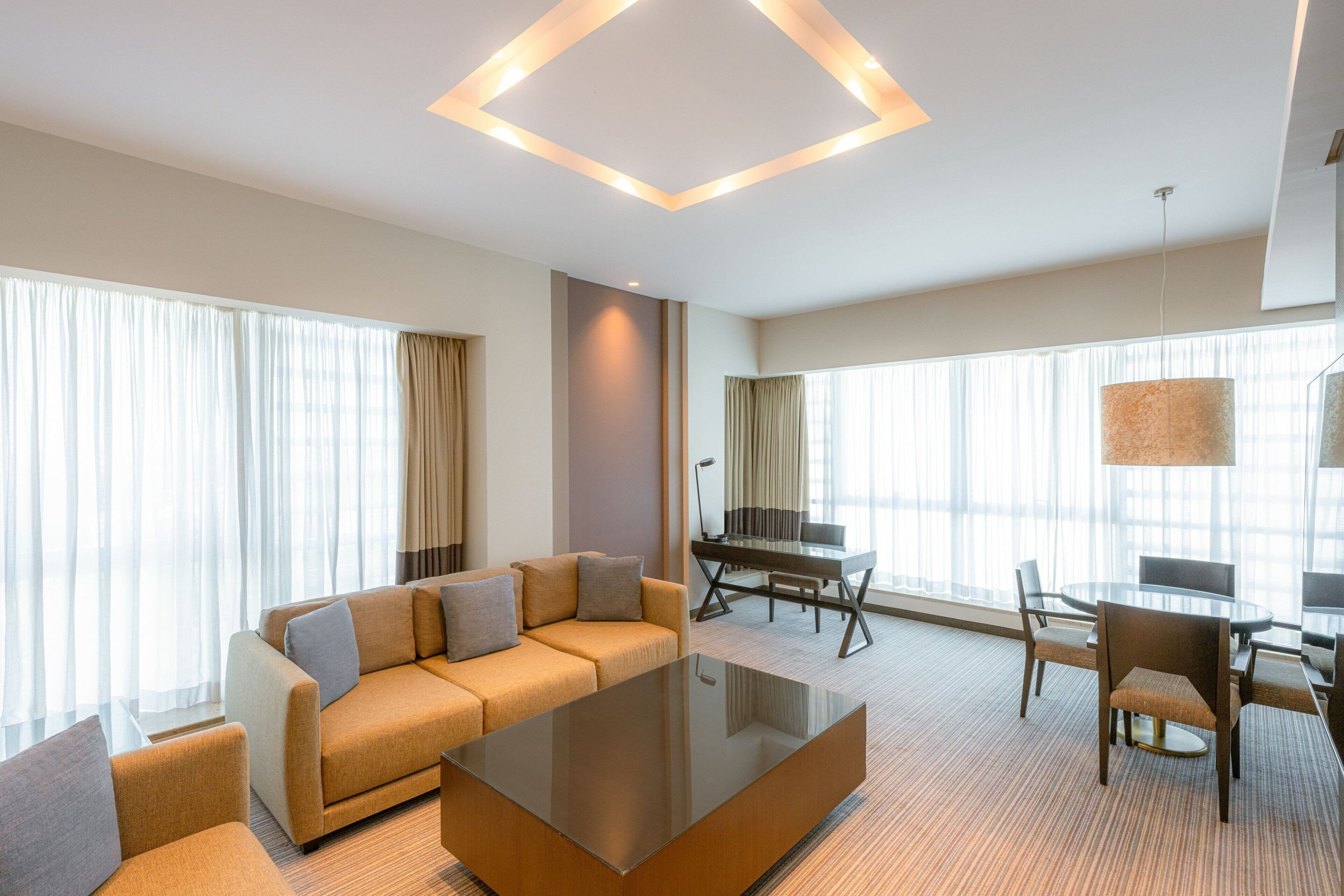 A modern hotel room with a sofa, a glass coffee table, a dining table with chairs, and large windows with curtains.