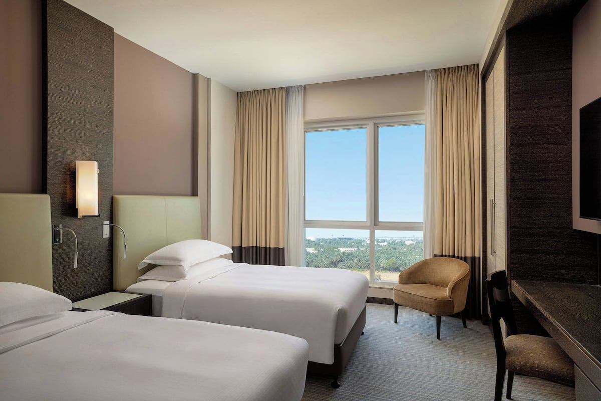 A hotel room with two beds, a desk, a chair, and a large window offering a city view.