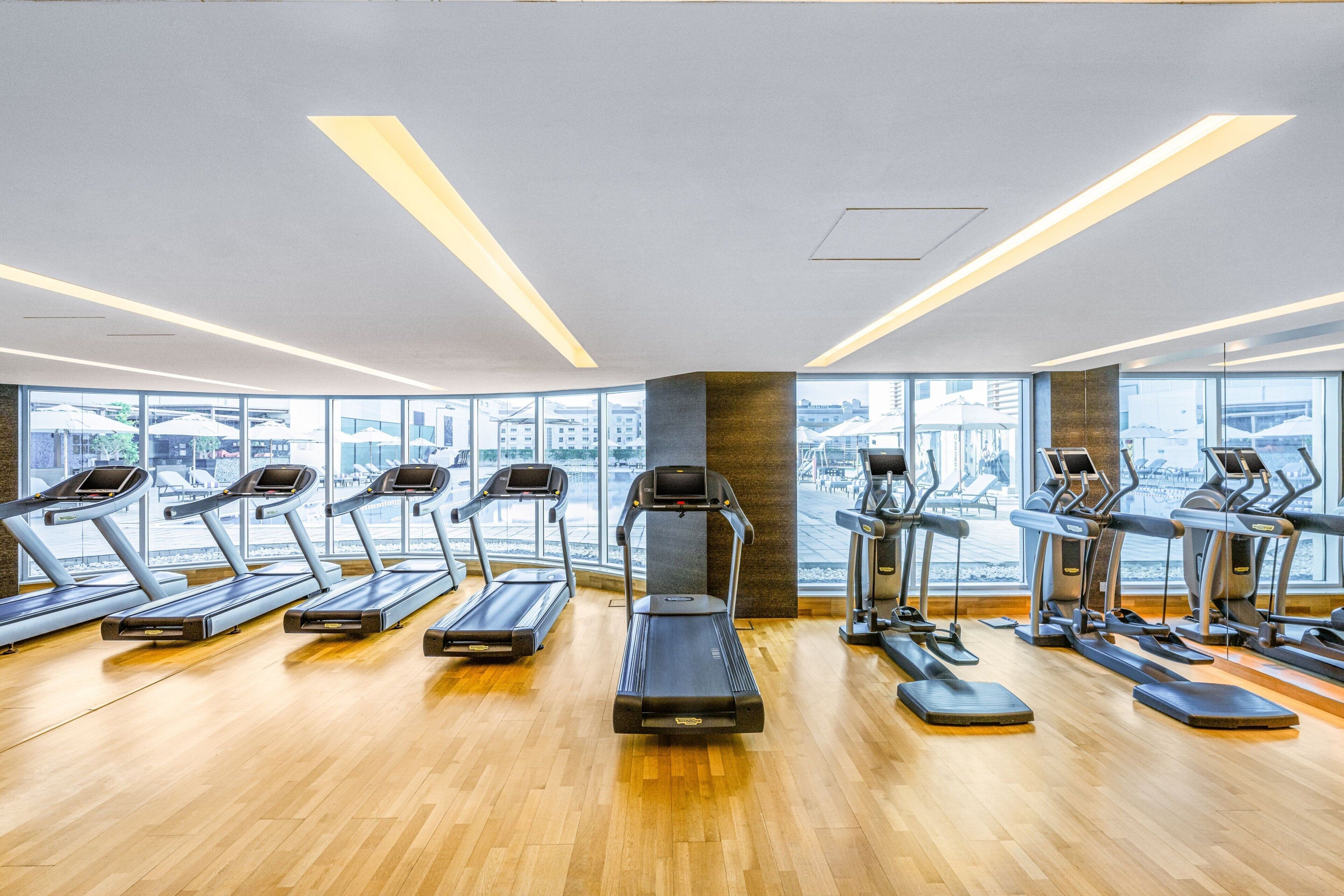 A modern gym with multiple treadmills and exercise equipment, large windows offering a view of the city, and a wooden floor.