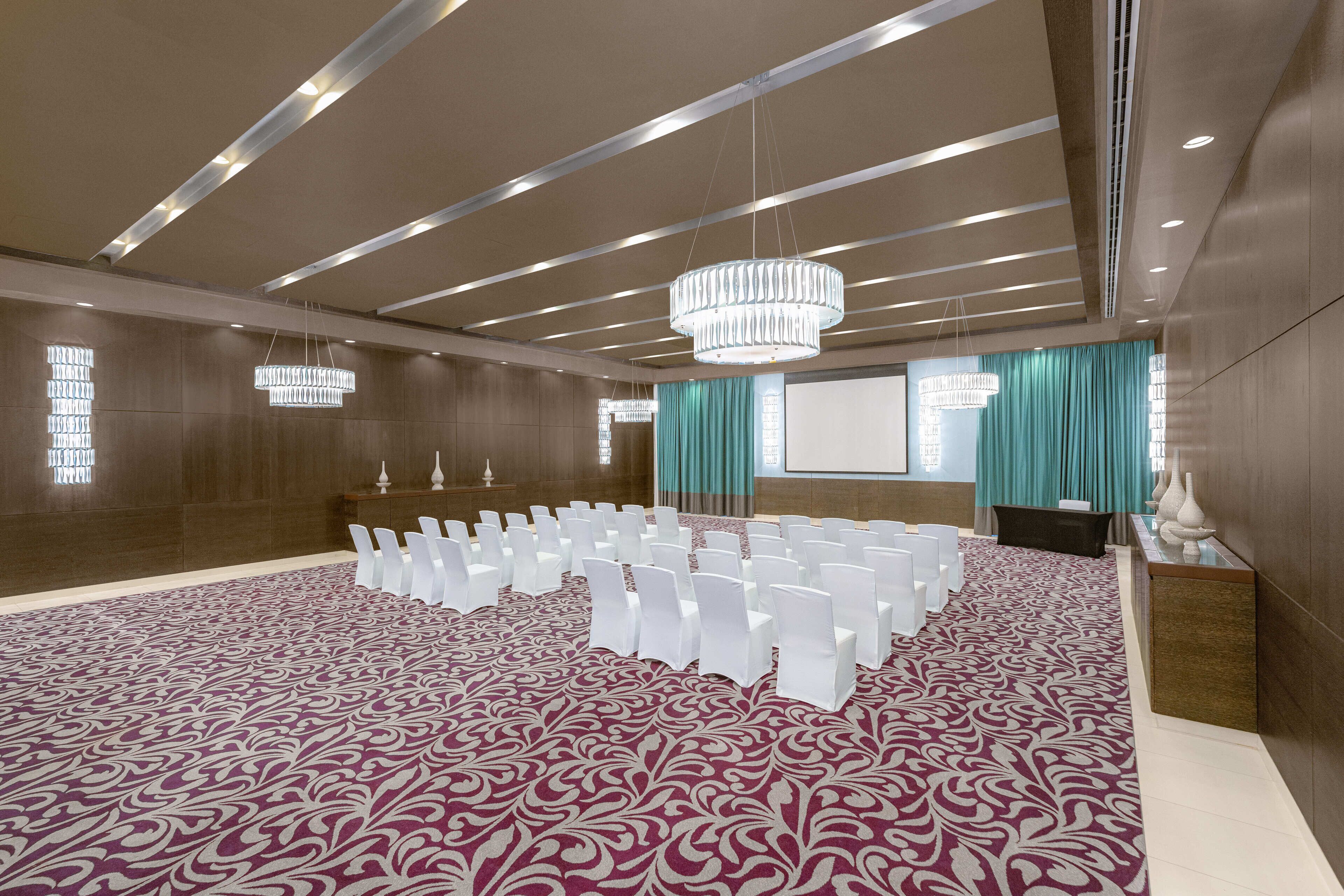 Un salón de conferencias con filas de sillas blancas, una lámpara de araña grande y una alfombra estampada.