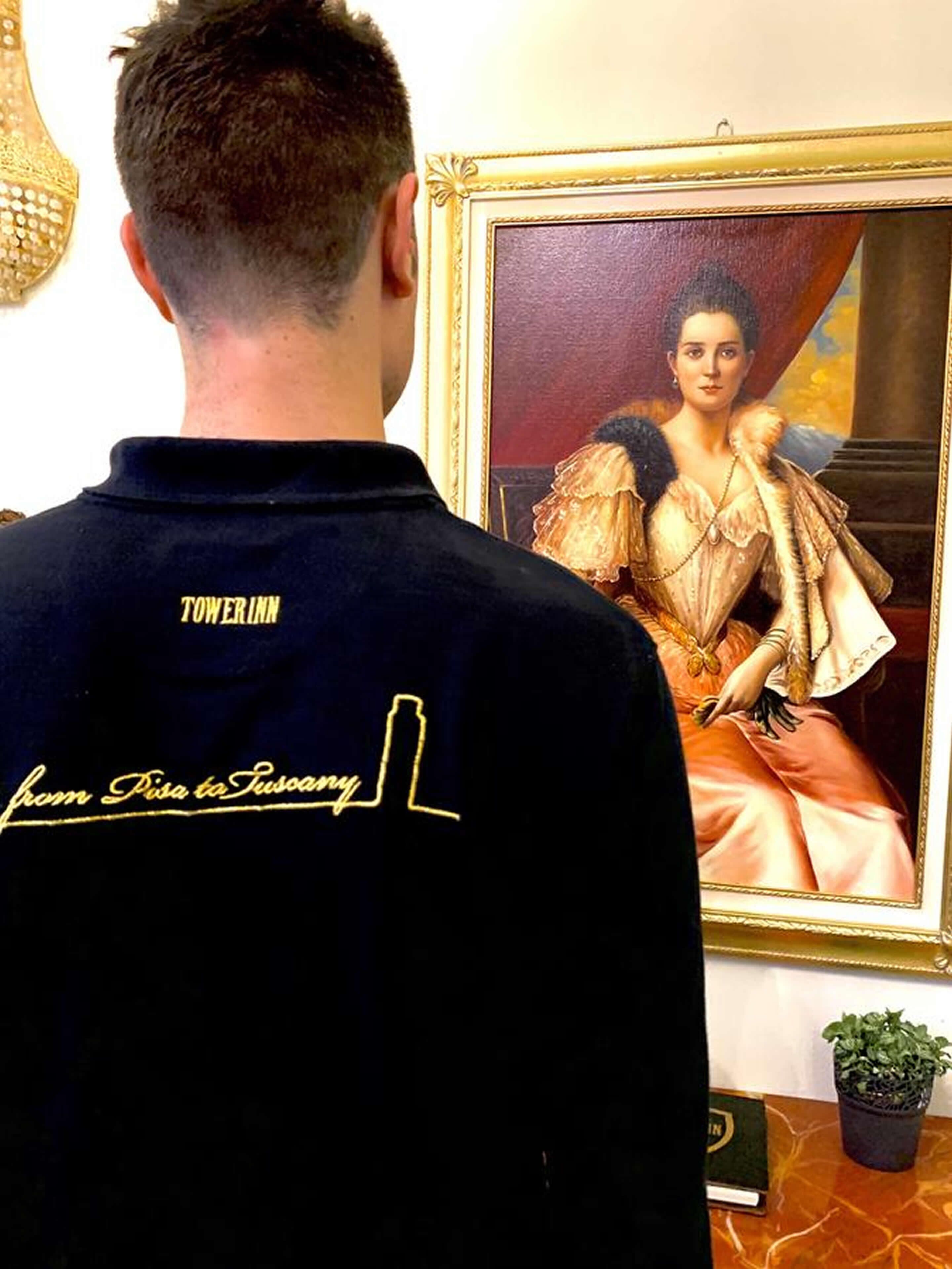 A person in a black jacket with "TOWERINN" and "from Pisa to Tuscany" text, standing in front of a framed painting.
