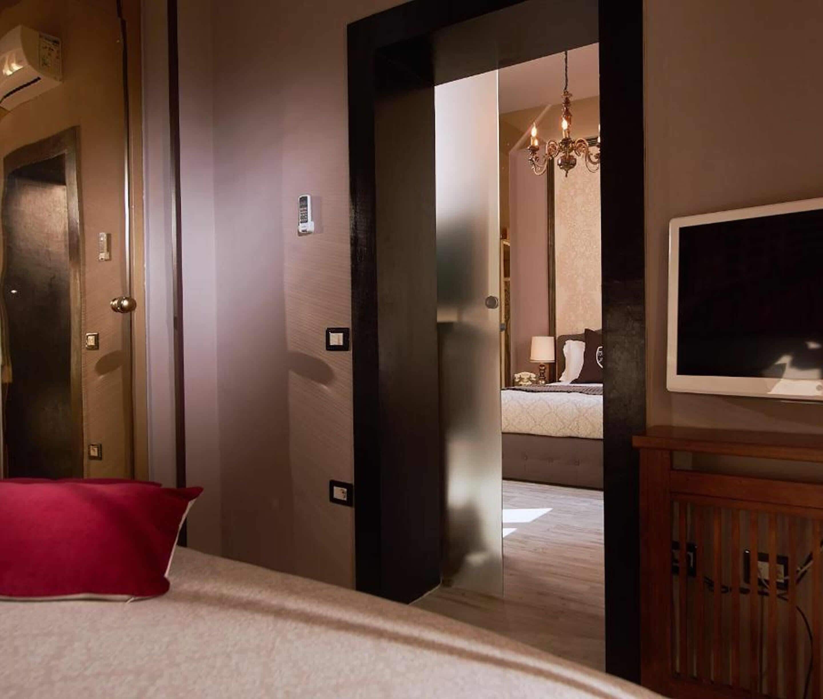 Una camera d'albergo con un letto, un cuscino rosso, una televisione e un lampadario sullo sfondo.