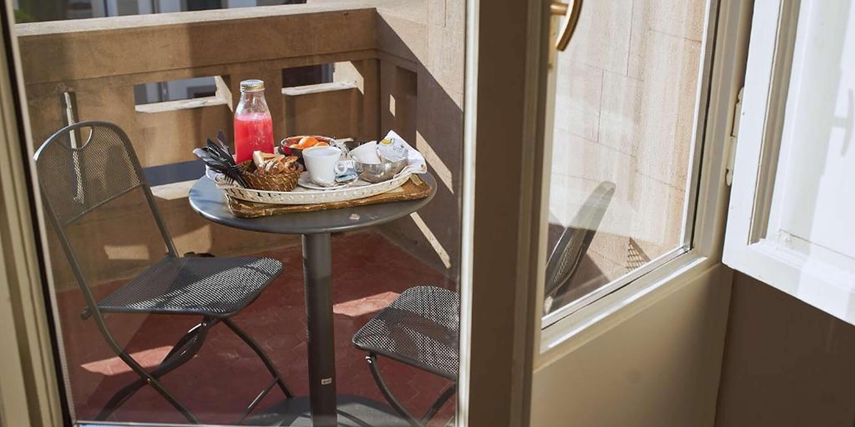 A small balcony with a table set for breakfast, including a basket of bread, a bottle of juice, and a cup.