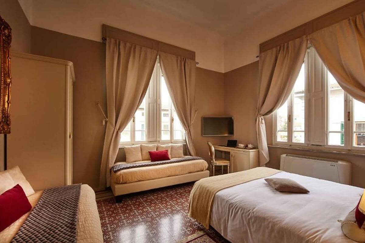 Habitación de hotel con cama, sofá, escritorio, televisor y ventanas amplias con cortinas.