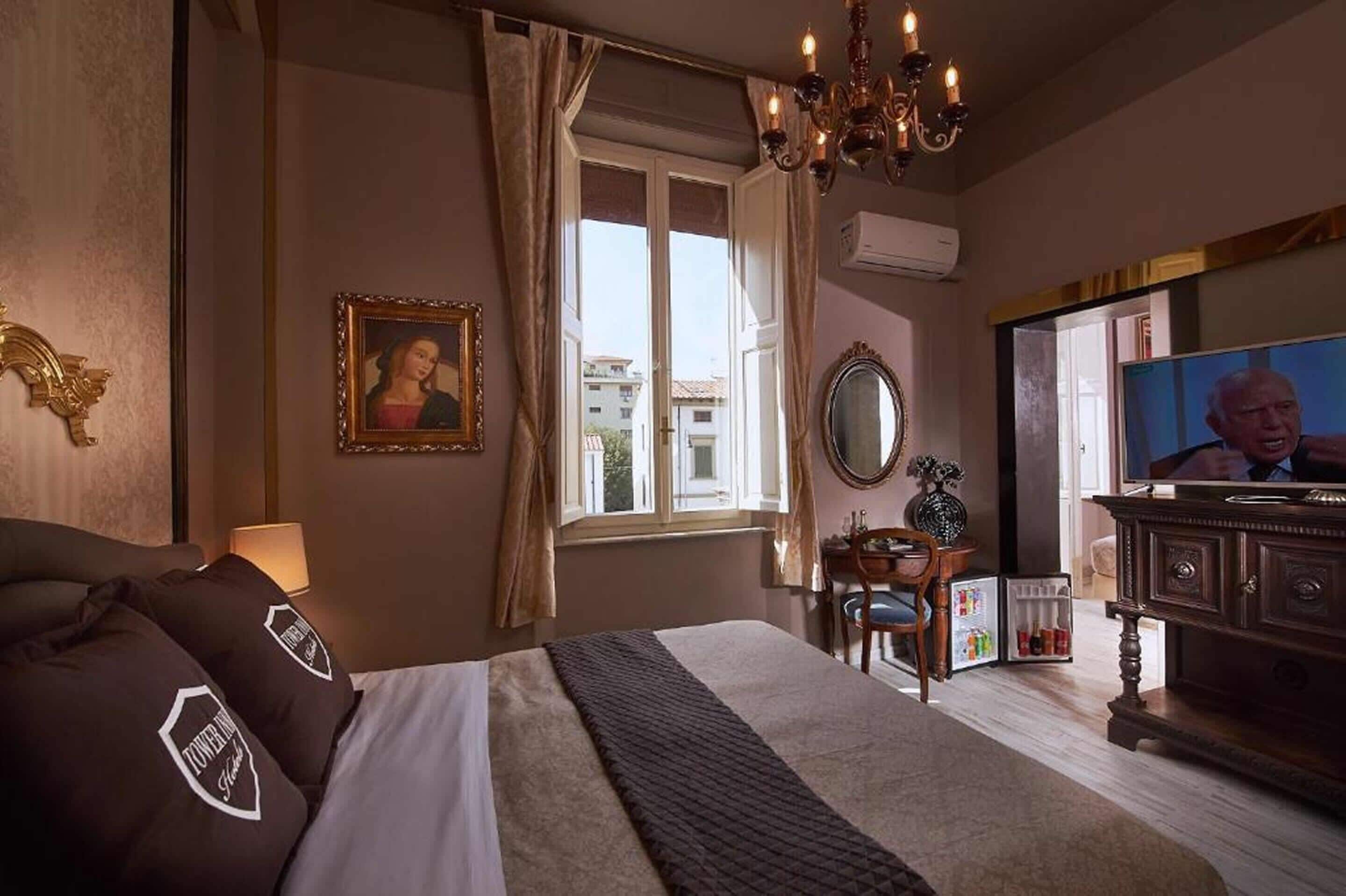 A hotel room with a bed, a television, a desk with a chair, a window with curtains, and a chandelier.