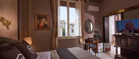 A hotel room with a bed, a television, a desk with a chair, a window with curtains, and a chandelier.